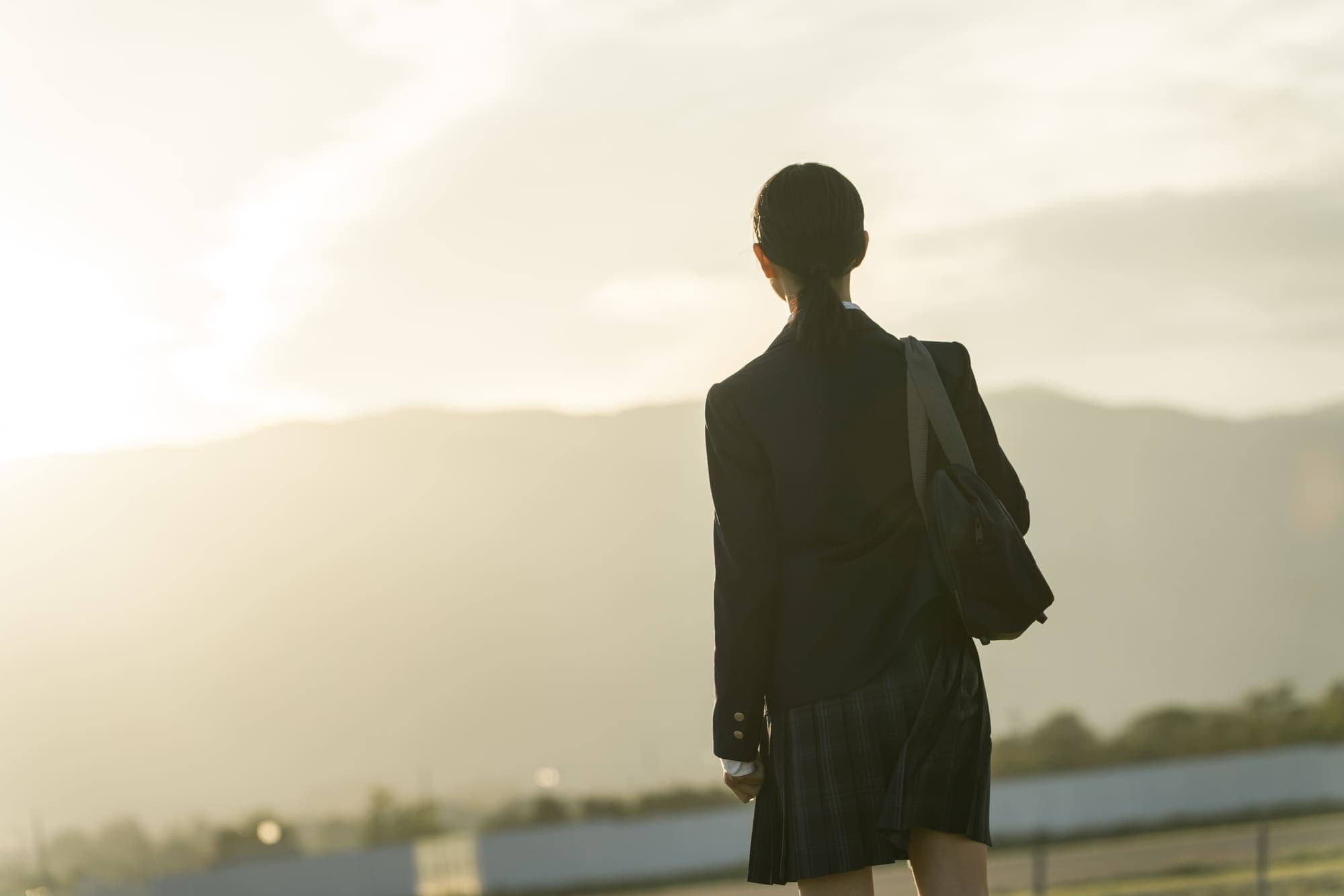 夕日の中、学校の制服で遠くを見つめる生徒の後ろ姿